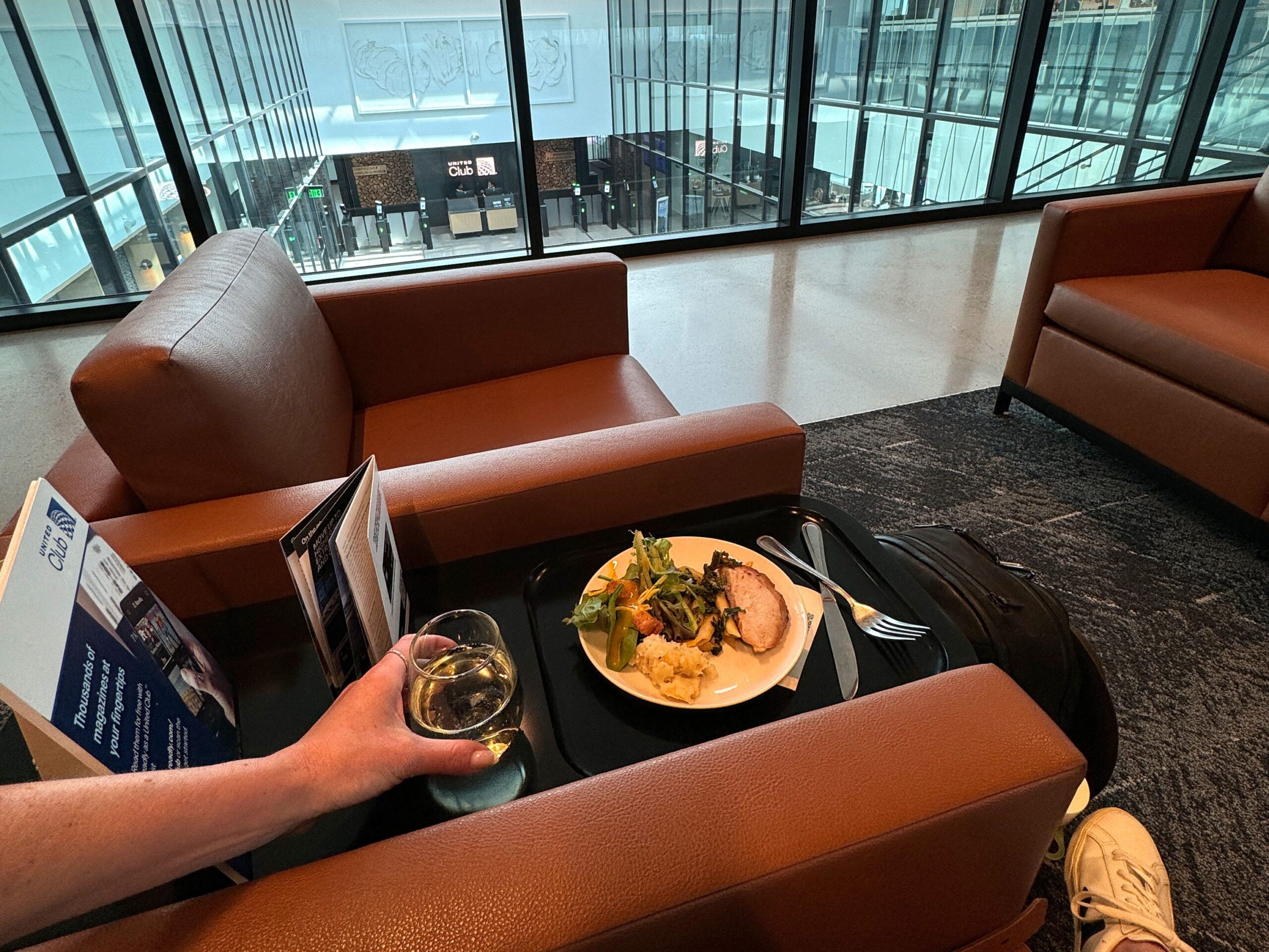 Emily sitting in a brown-leather chair, holding a glass of white wine with a plate of food on a table next to her. The walls are glass and see-through.