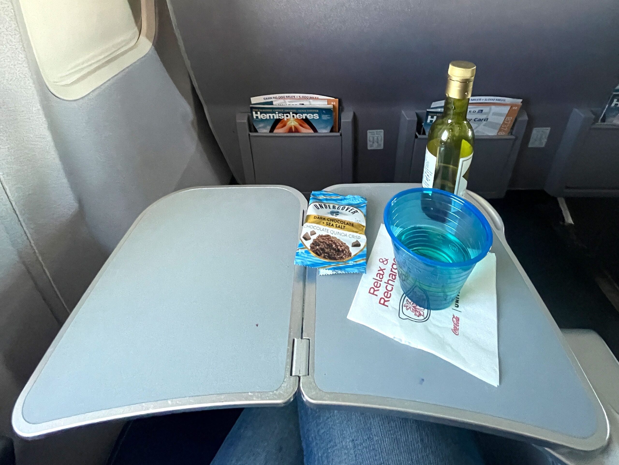 A cookie in a package, a small bottle of white wine, a blue cup, and a napkin sit on a tray on a plane.