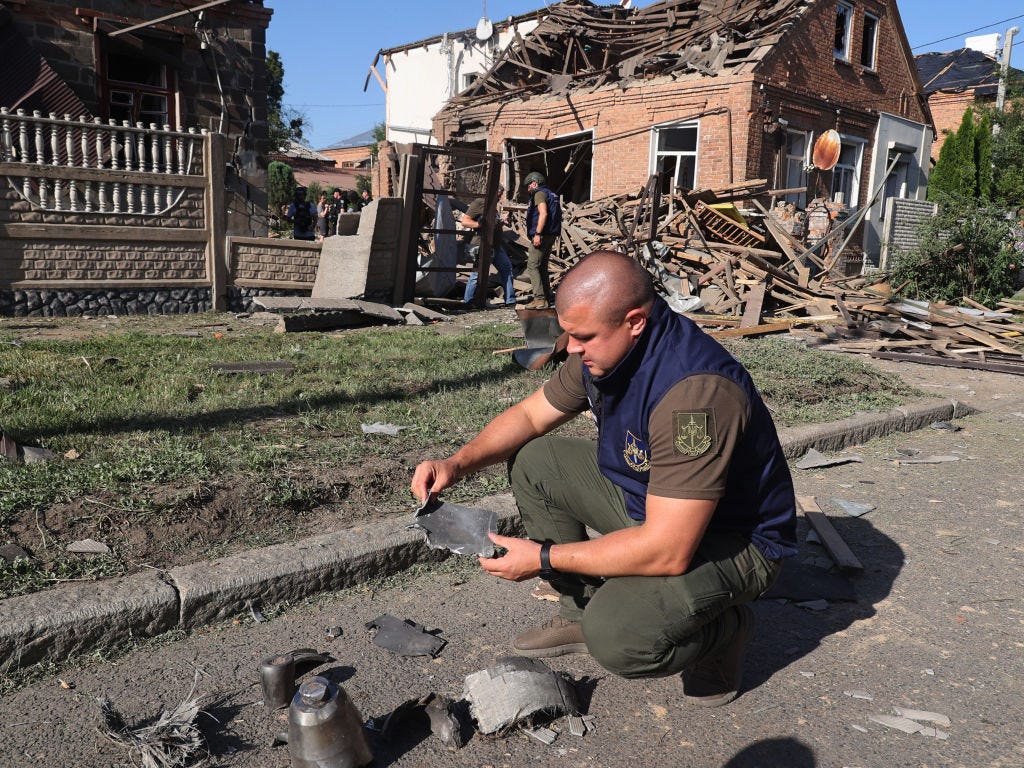 A Ukrainian Prosecutor's Office employee squatting down to analyze Russian munition fragments with destroyed homes in the background.