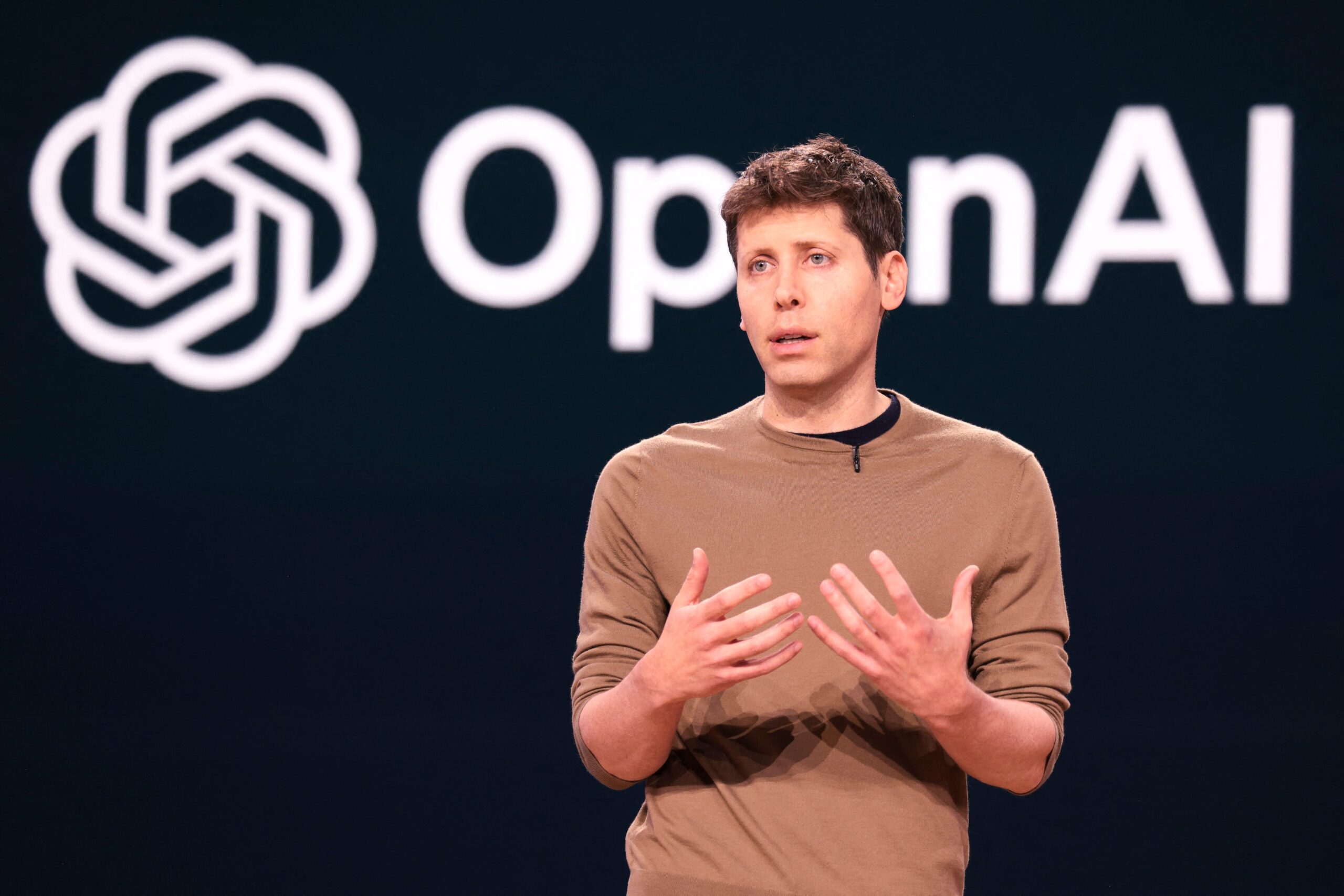 Sam Altman presenting onstage with the OpenAI logo behind him.