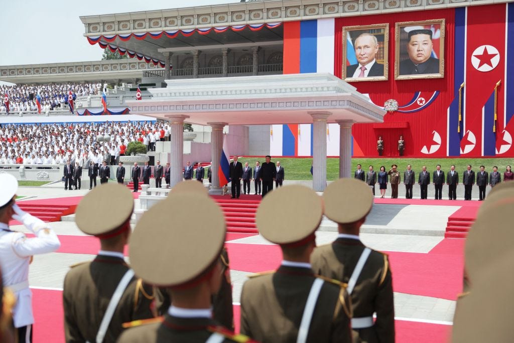 Kim Jong Un and Vladimir Putin stand under a stone gazebo surrounded by people in military dress and others in suits.