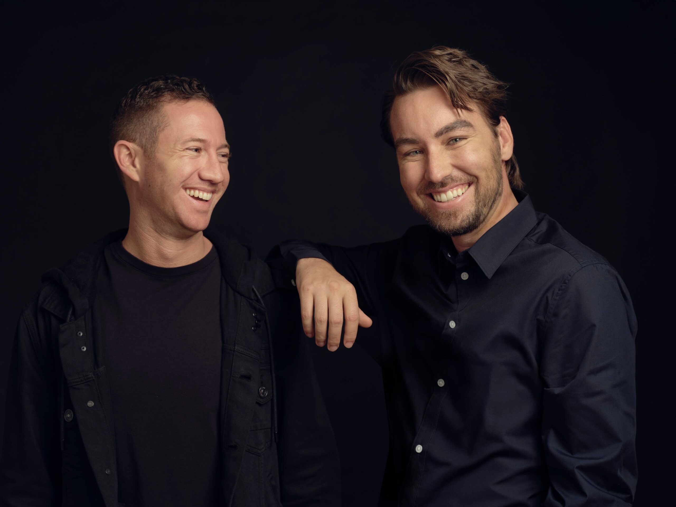 Two men looking relaxed smile for a photo against a black backdrop.